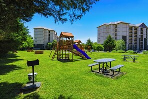 Um parquinho com escorrega e estruturas de escalada, cercado por um gramado bem cuidado, mesas de piquenique e um edifício ao fundo.