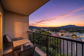 A balcony with a table and chairs, overlooking a parking lot and a town with mountains in the background.