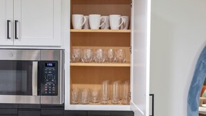 Una cocina con microondas, un reloj digital y un mueble con estantes que contienen tazas blancas y vasos de vidrio.
