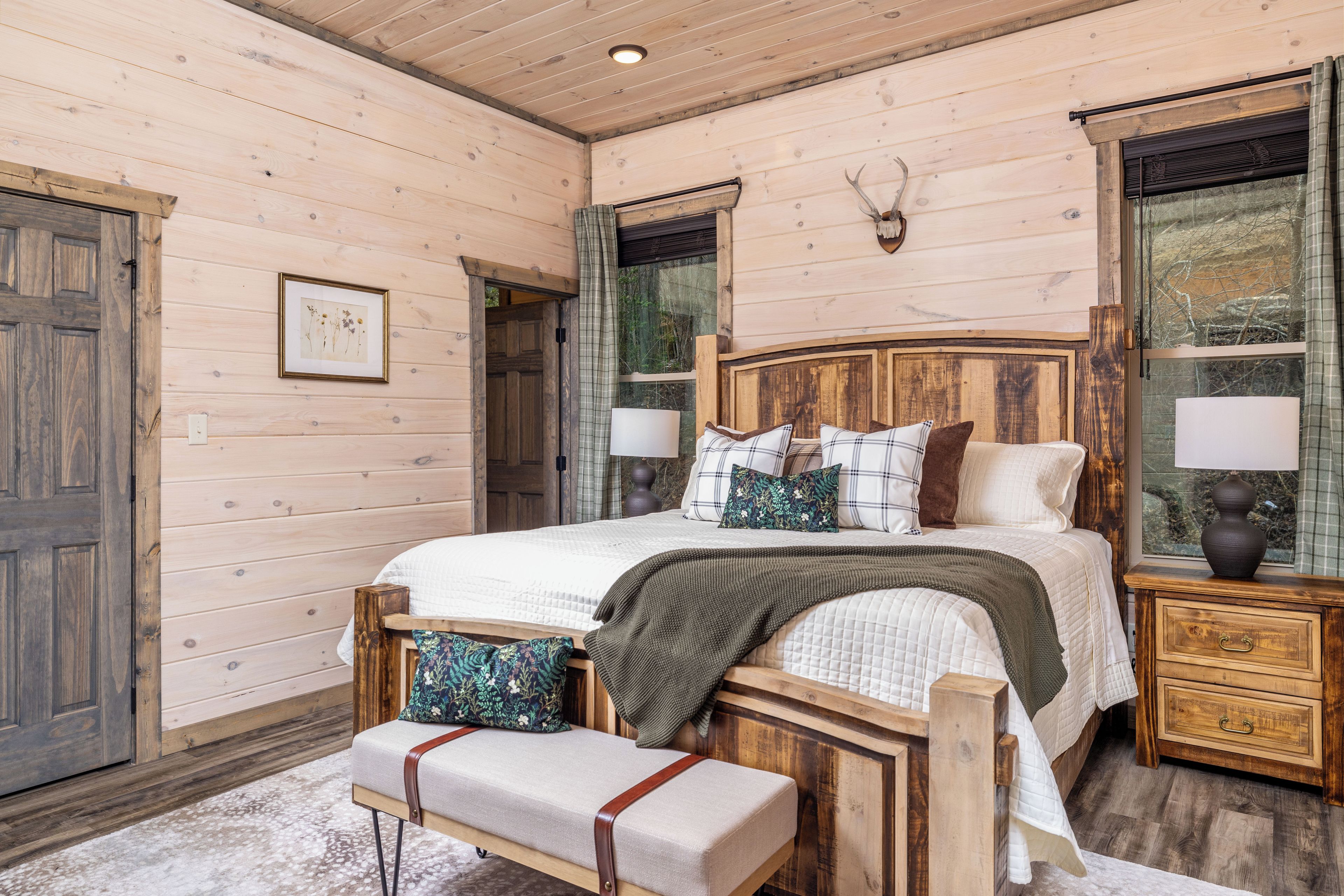 Une chambre à coucher aux murs lambrissés, avec un lit, un banc, des tables de chevet et une tête de cerf accrochée au mur.