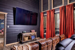 A room with a large flat-screen TV mounted on the wall, a wooden entertainment unit with storage, leather sofas, and red curtains on the windows. - Mountain Explorers Retreat (Gatlinburg)
