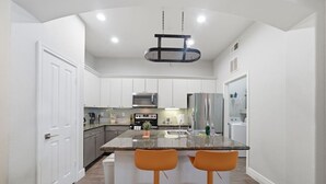 A modern kitchen with white cabinetry, a granite island, and two orange bar stools. - Sleeps 5 | Downtown & Sport Stadiums Nearby | Pool + Gym + Free Parking (Houston)