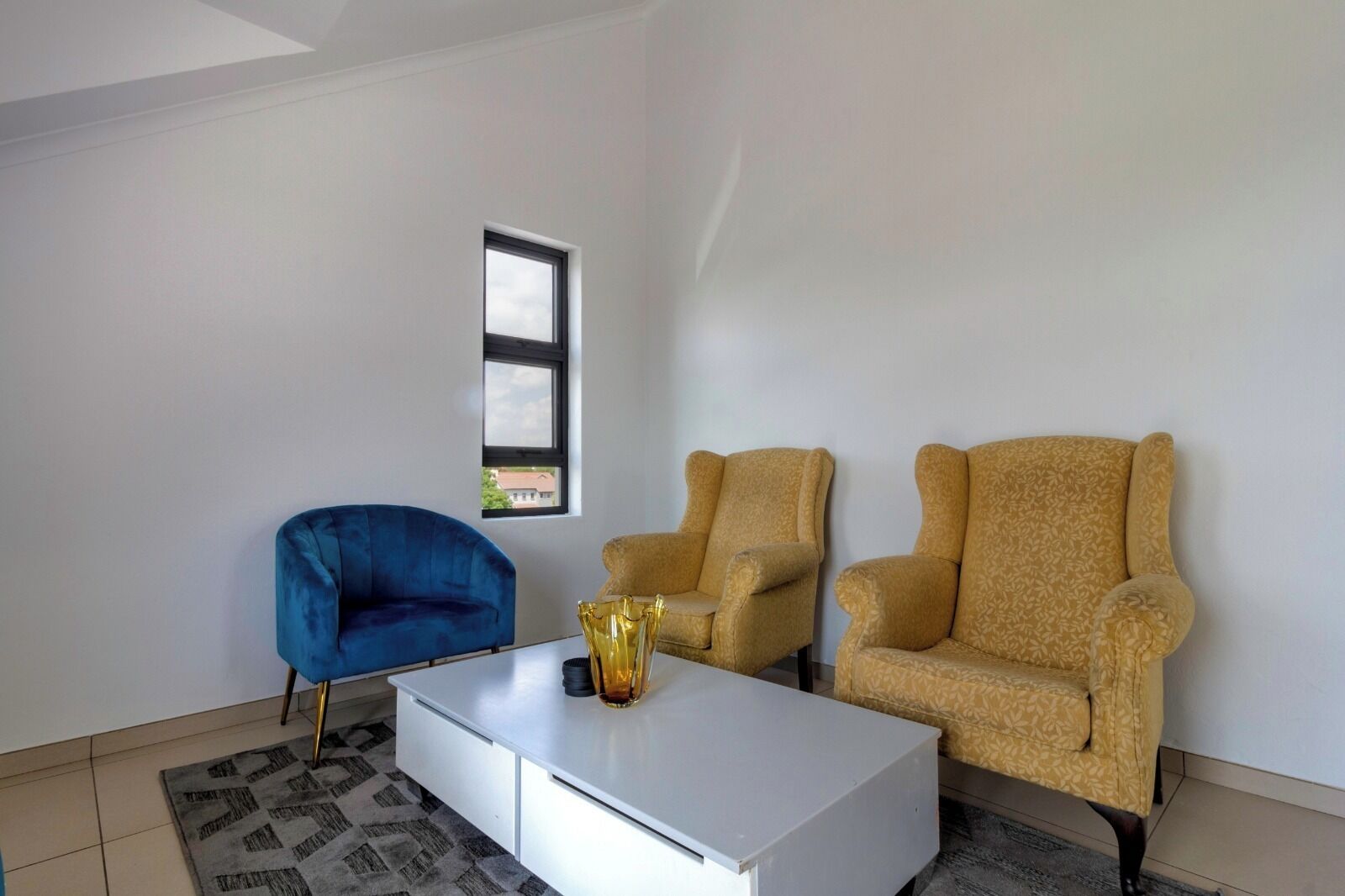 A modern living room with a white sofa, a blue armchair, and two patterned armchairs. A white coffee table with a gold vase is in the center.