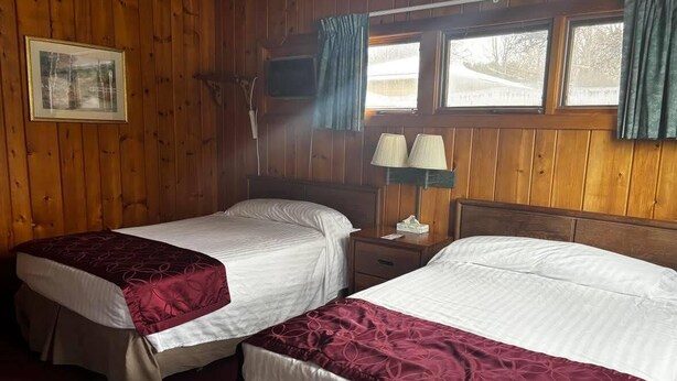 Une chambre avec deux lits, des murs en bois, un tableau encadré, une lampe et une fenêtre avec des rideaux.