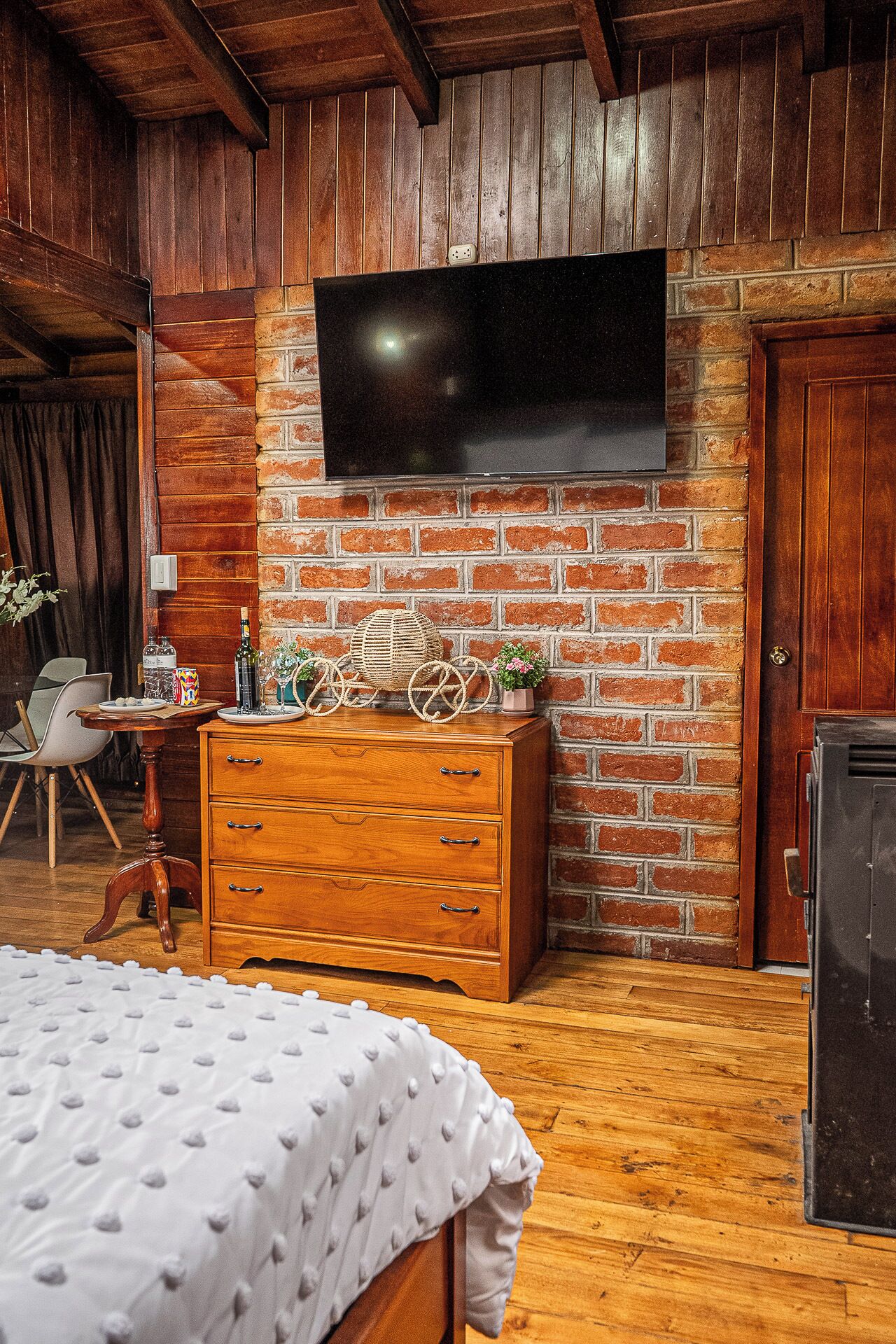 Quarto com parede de tijolos, uma televisão de tela plana fixada acima de uma cômoda de madeira, cama com colcha branca e piso de madeira.