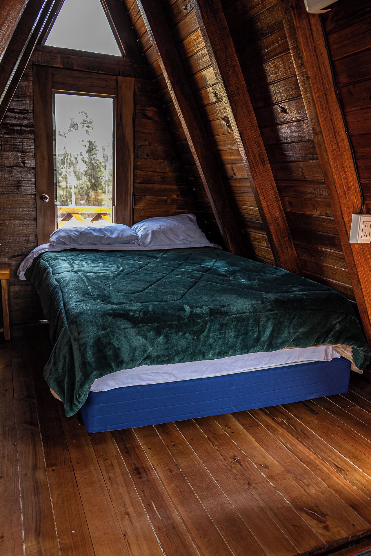 Una habitación con techo inclinado, paredes de madera, una cama con cubrecama verde y una ventana con vistas a árboles.