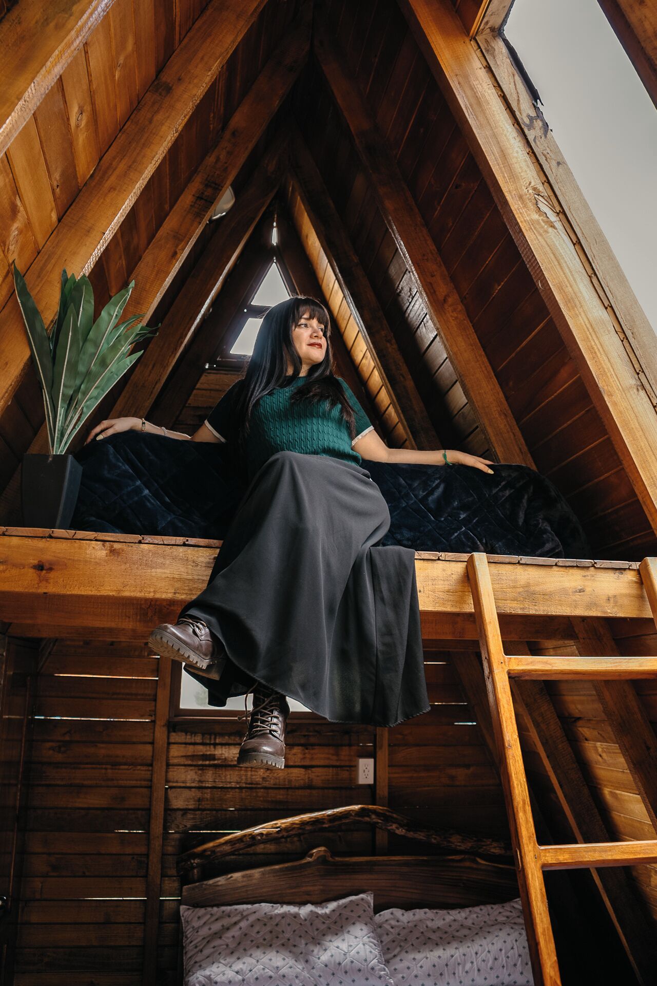 Una persona sentada en una escalera de madera en un altillo con techo inclinado, una ventana y una planta.