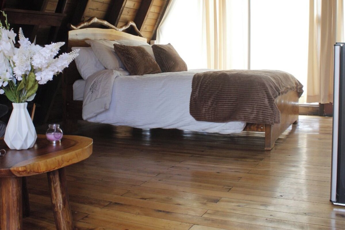 Un dormitorio tipo loft con techo inclinado de madera, una cama con ropa de cama blanca y un plaid marrón, una mesa de madera con un jarrón de flores y un ventanal con cortinas.