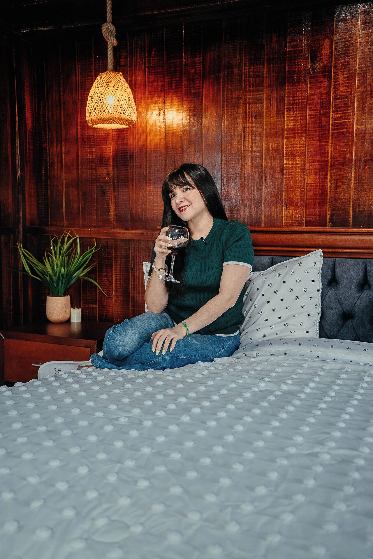 A person sitting on a bed with a patterned bedspread, holding a glass.