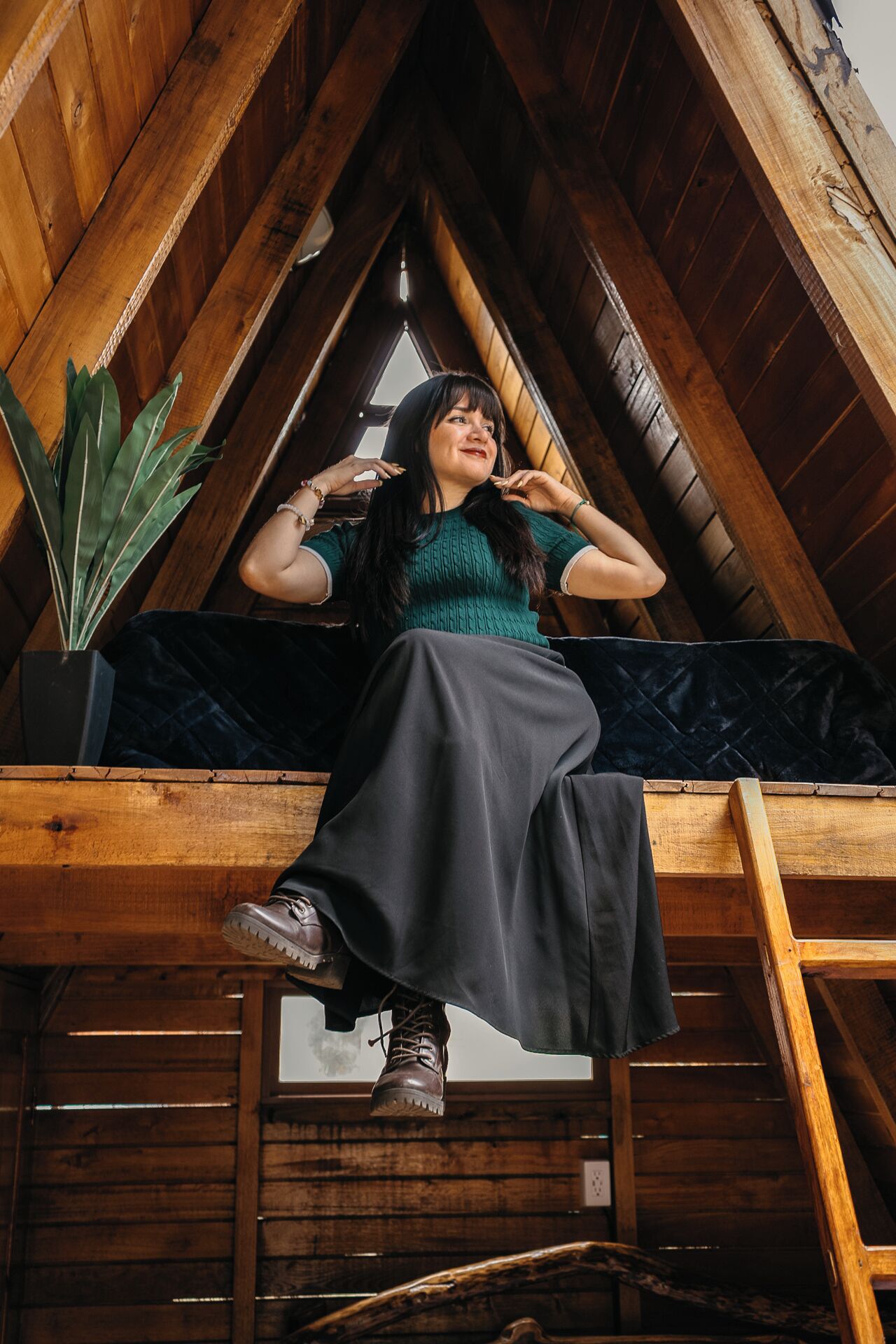 Una persona sentada sobre una viga de madera en una habitación con techo inclinado, una planta en maceta y una ventana.