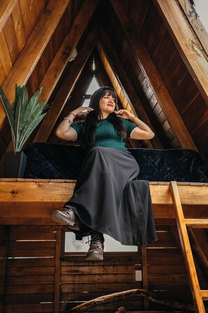 Una persona sentada sobre una viga de madera en una habitación con techo inclinado, una planta en maceta y una ventana.