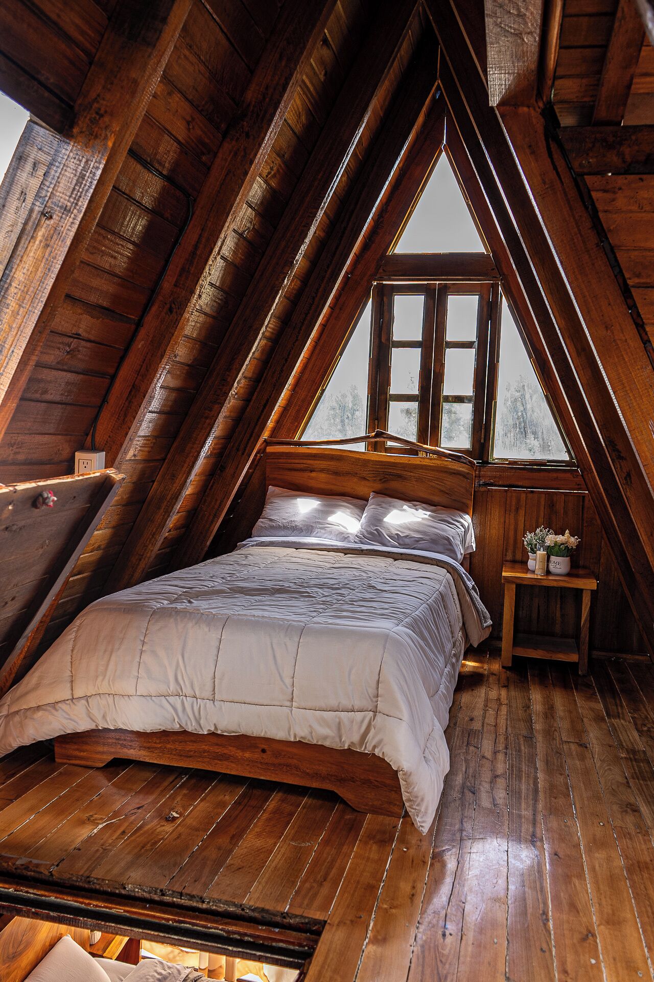 Une chambre à coucher aménagée dans un espace mansardé, avec un plafond en pente, des poutres en bois, un lit recouvert d’une couette blanche, une table de chevet ornée d’un vase et une fenêtre donnant sur la forêt.