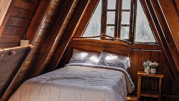 Une chambre à coucher aménagée dans un espace mansardé, avec un plafond en pente, des poutres en bois, un lit recouvert d’une couette blanche, une table de chevet ornée d’un vase et une fenêtre donnant sur la forêt.