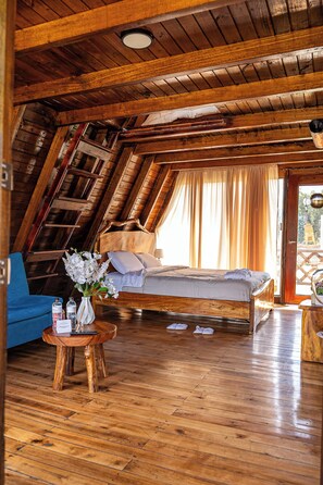 Une chambre mansardée avec un plafond en bois, un lit, une chaise bleue et une petite table sur laquelle est posé un vase de fleurs.