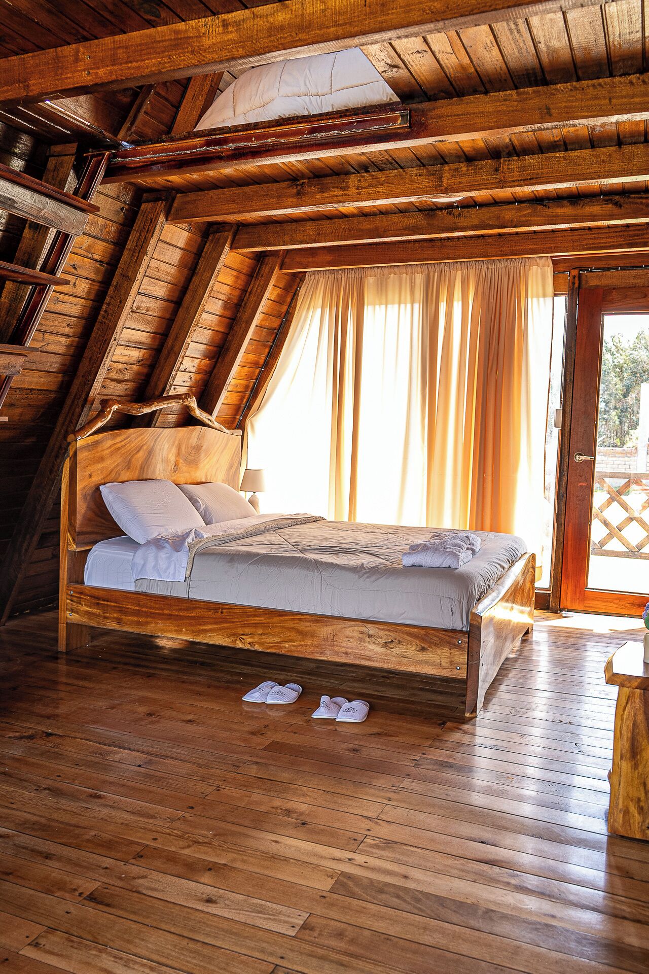 A wooden bed in a room with sloped ceilings and a view through a window.
