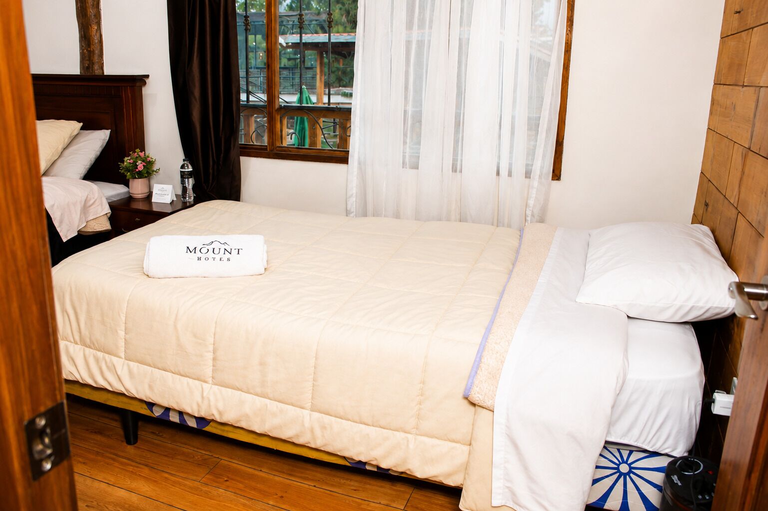 Una cama bien hecha con una manta blanca y una toalla doblada con la marca 'MOUNT HOTELS', un cabecero de madera, una mesita de noche con una lámpara y una ventana con cortinas.