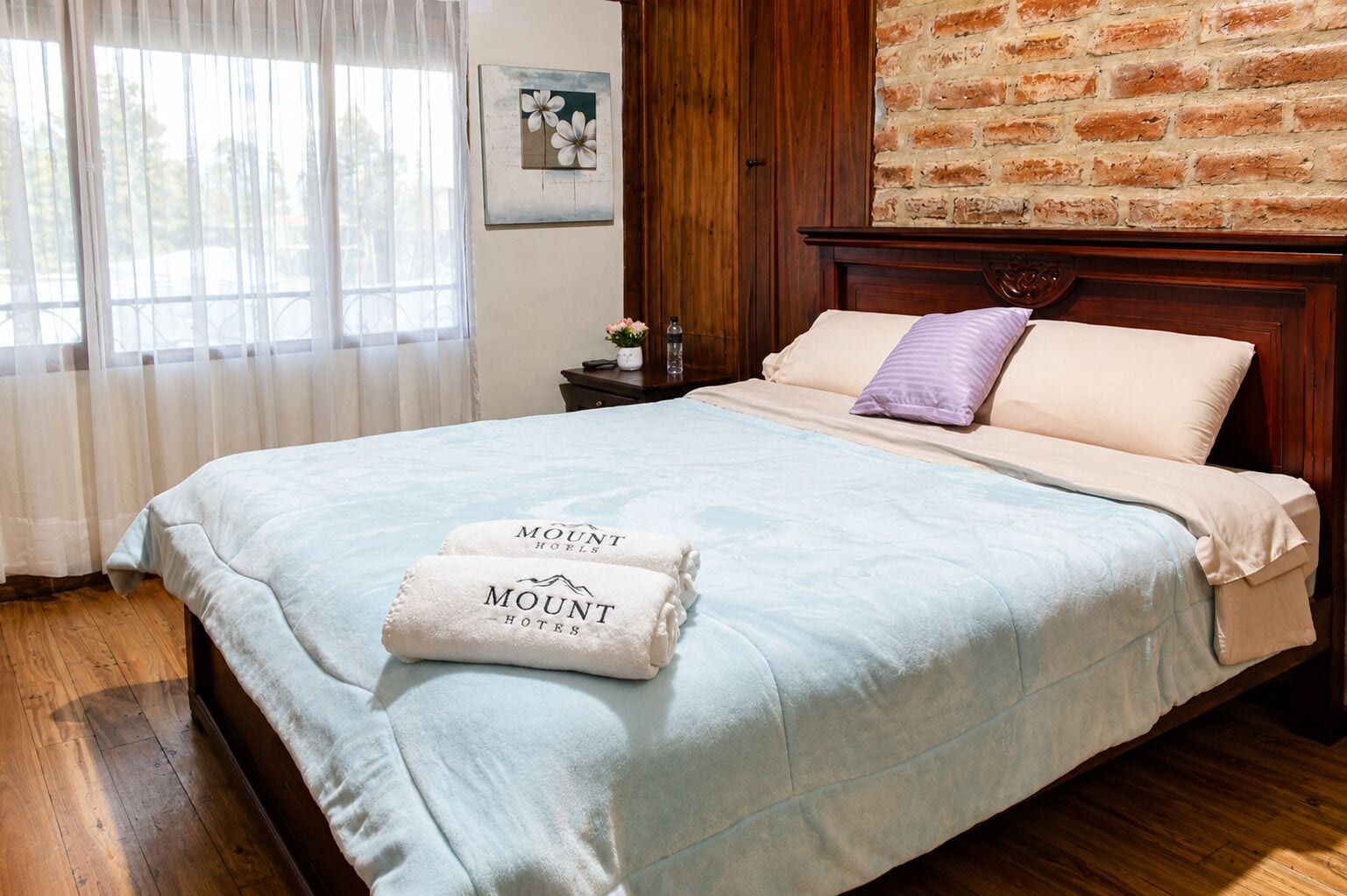 Habitación de hotel con cabecera de madera, cama con colcha azul claro y dos toallas con el nombre del hotel.