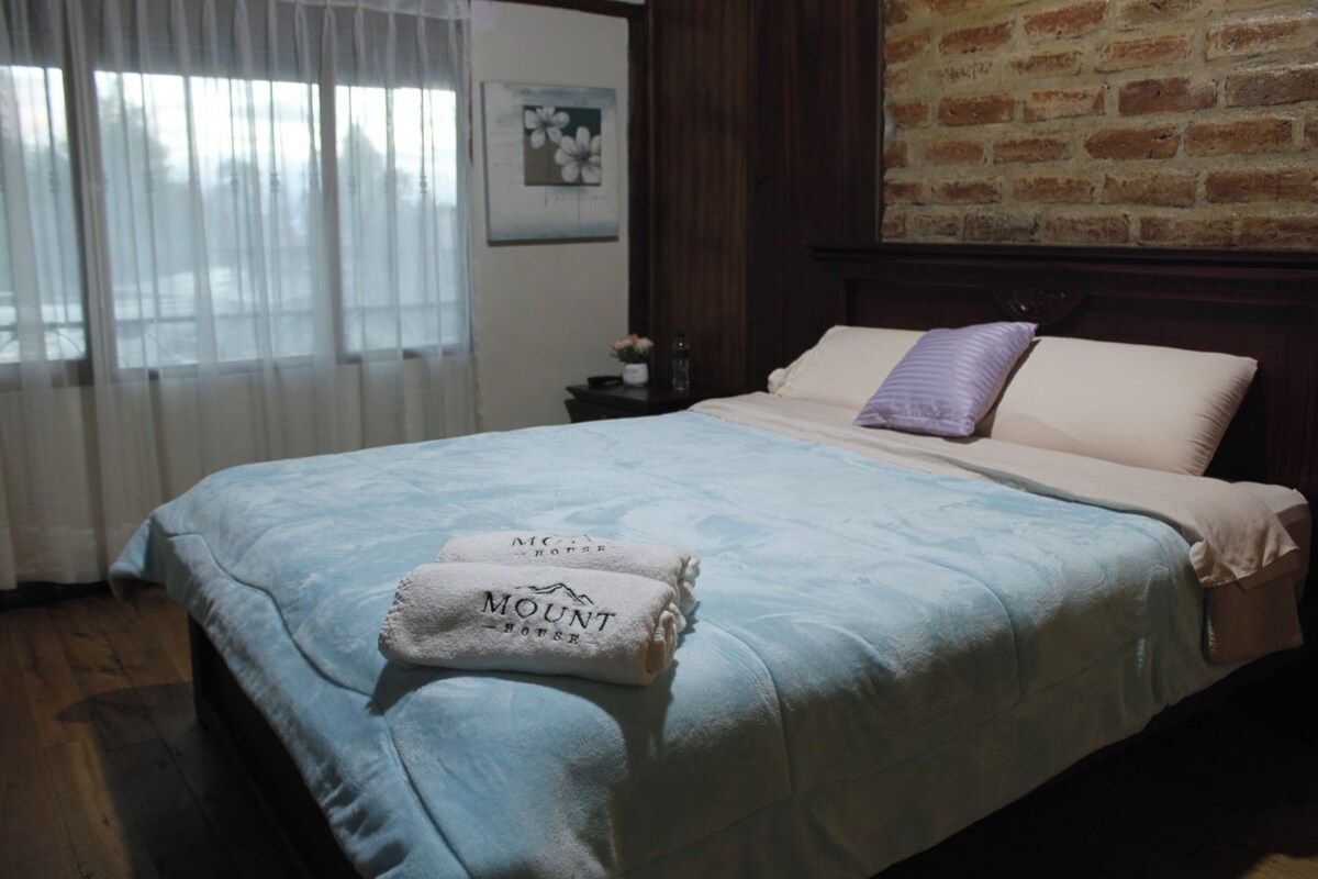 Una habitación de hotel con cama, mesita de noche, pared de ladrillo y ventana con cortinas.