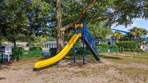 A playground with a yellow slide and blue structures, surrounded by trees and a fence. - Newly remodeled 3BR/2BA Oceanfront Golf Cart Village Beach House (Surfside Beach)
