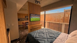 Ein Schlafzimmer mit einem Bett, einem an einer Holzwand montierten Fernseher, einem Holztisch und Blick auf einen umzäunten Außenbereich.