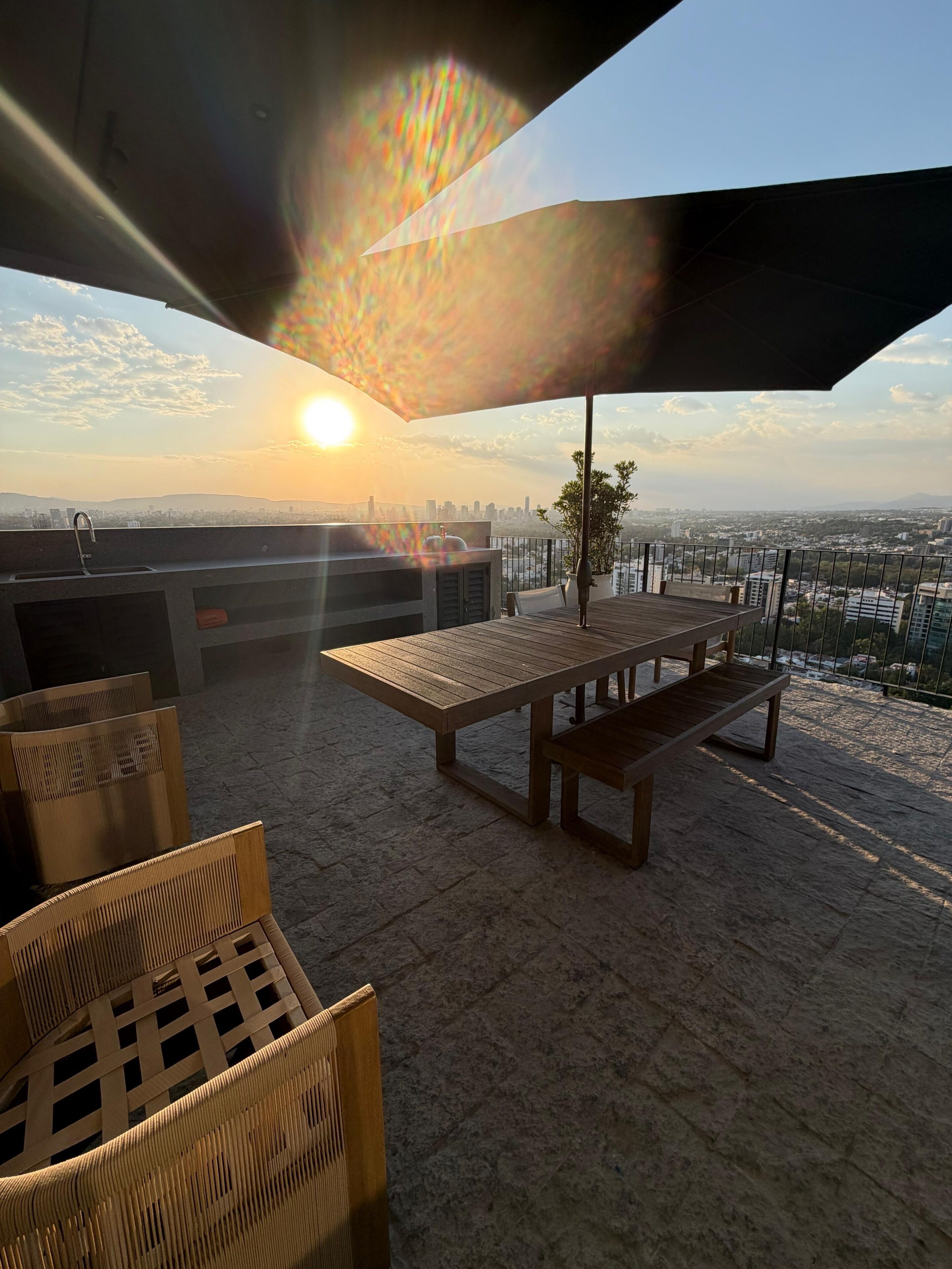 Une terrasse extérieure avec une table et des chaises, un barbecue et une vue sur les toits d’une ville au coucher du soleil.