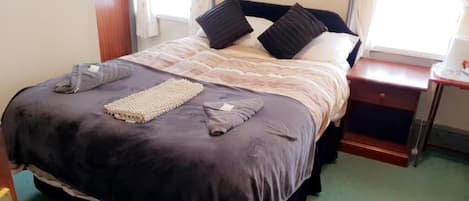 A neatly made bed with a patterned bedspread and pillows, a bedside table with a lamp, and a window with curtains.