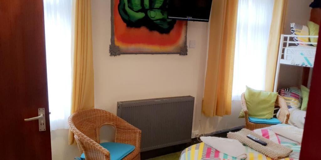 A room with a wicker chair, a bed with a colorful bedspread, a radiator, and a wall-mounted television.