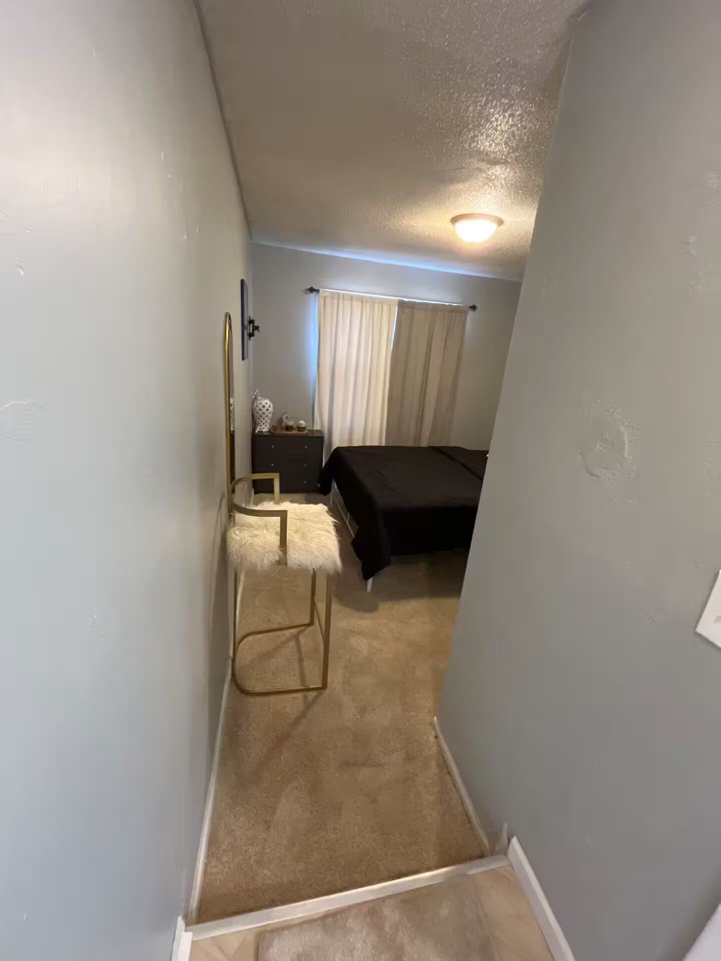 A narrow hallway leading to a bedroom with a bed, a nightstand, and a chair.