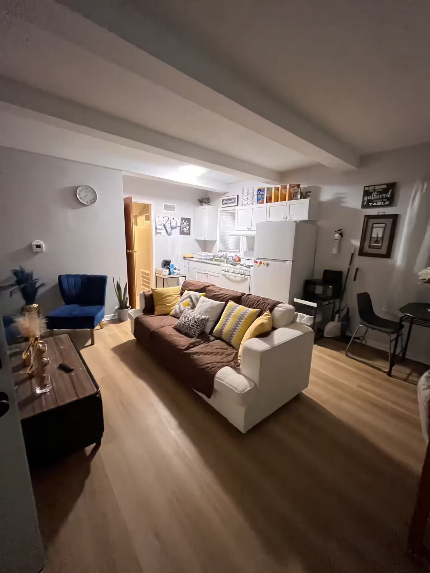 A living room with a brown sofa, a blue chair, a small table, and a kitchen area in the background.