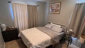 Un dormitorio con cama, cortinas, una mesita de noche, una lámpara y un cuadro en la pared.