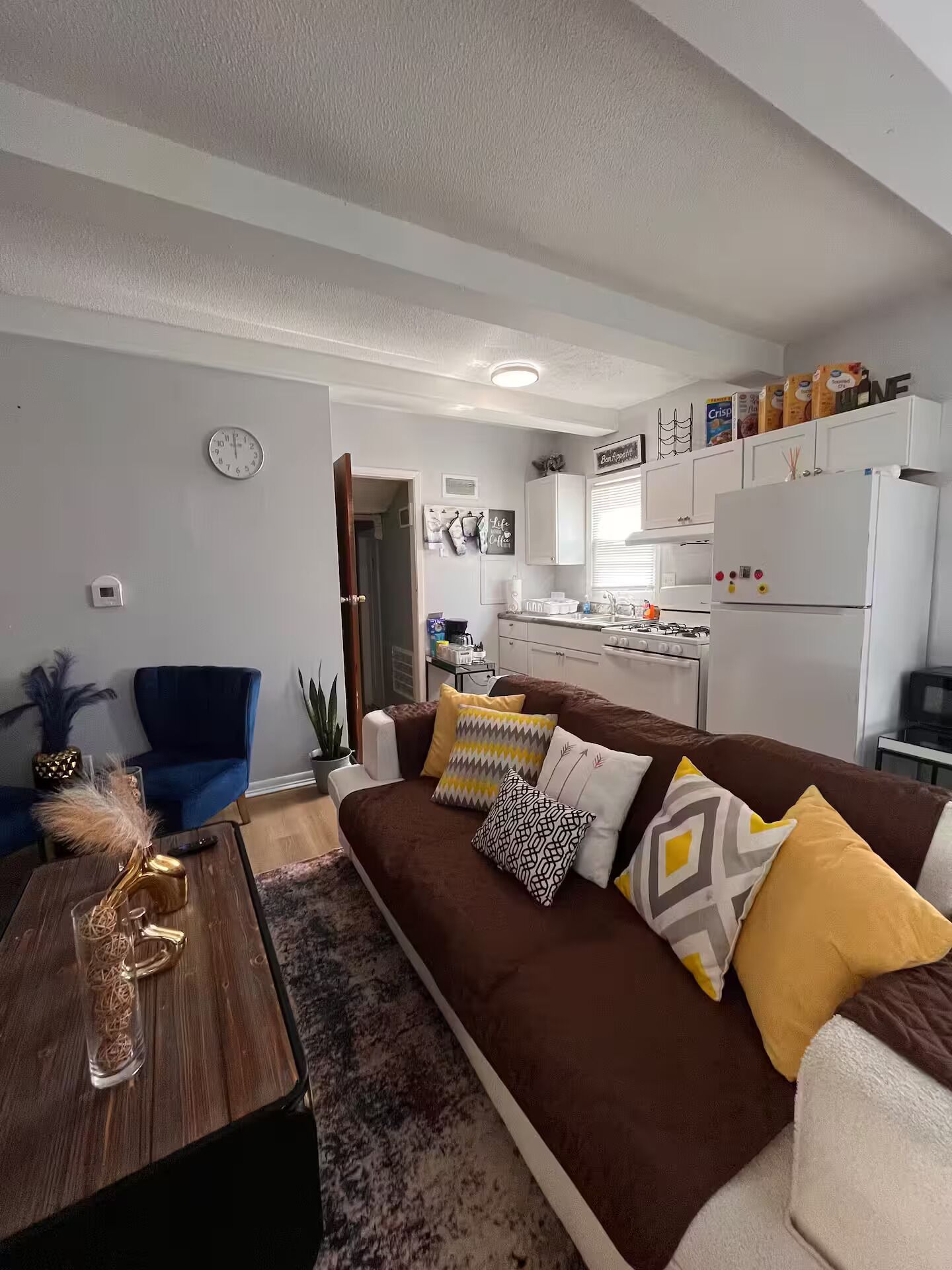 A living room with a brown sofa, a coffee table, and a kitchen area with white cabinets and appliances.