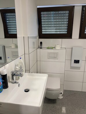 Ein Badezimmer mit weißem Waschbecken, Toilette, Spiegel und einem Fenster mit Jalousien.
