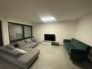 Ein Wohnzimmer mit grauem Sofa, grünem Sofa, Flachbildfernseher und Fenster mit Jalousien.