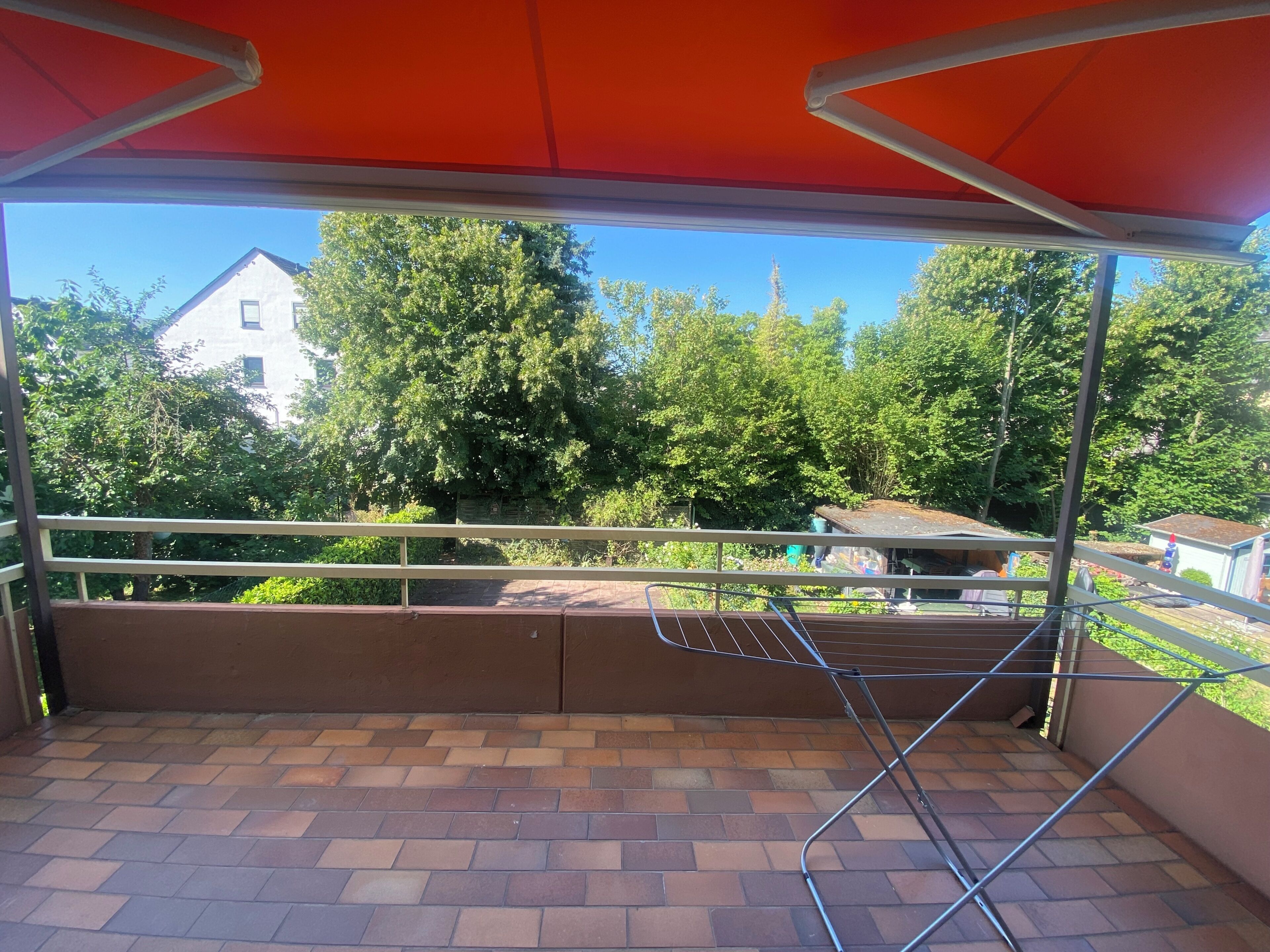 A covered balcony with a metal railing, brick flooring, and a view of trees and buildings in the background.