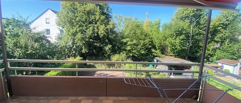 A covered balcony with a metal railing, brick flooring, and a view of trees and buildings in the background.