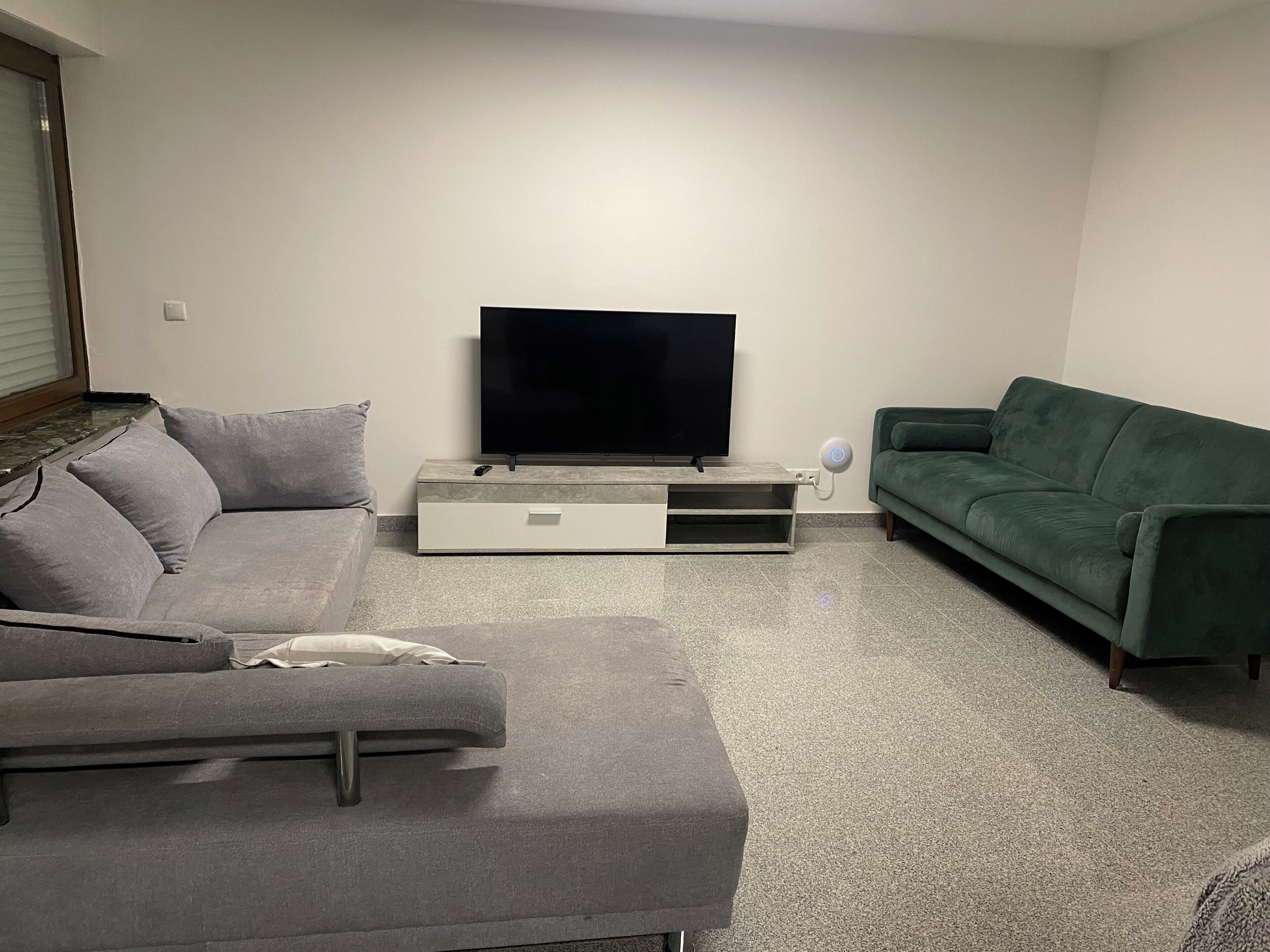Una sala de estar con un sofá gris, un sofá verde, un televisor de pantalla plana sobre un soporte y una pared blanca.