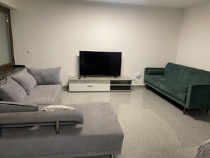 Ein Wohnzimmer mit grauem Sofa, grünem Sofa, Flachbildfernseher auf einem Schrank und einer weißen Wand.