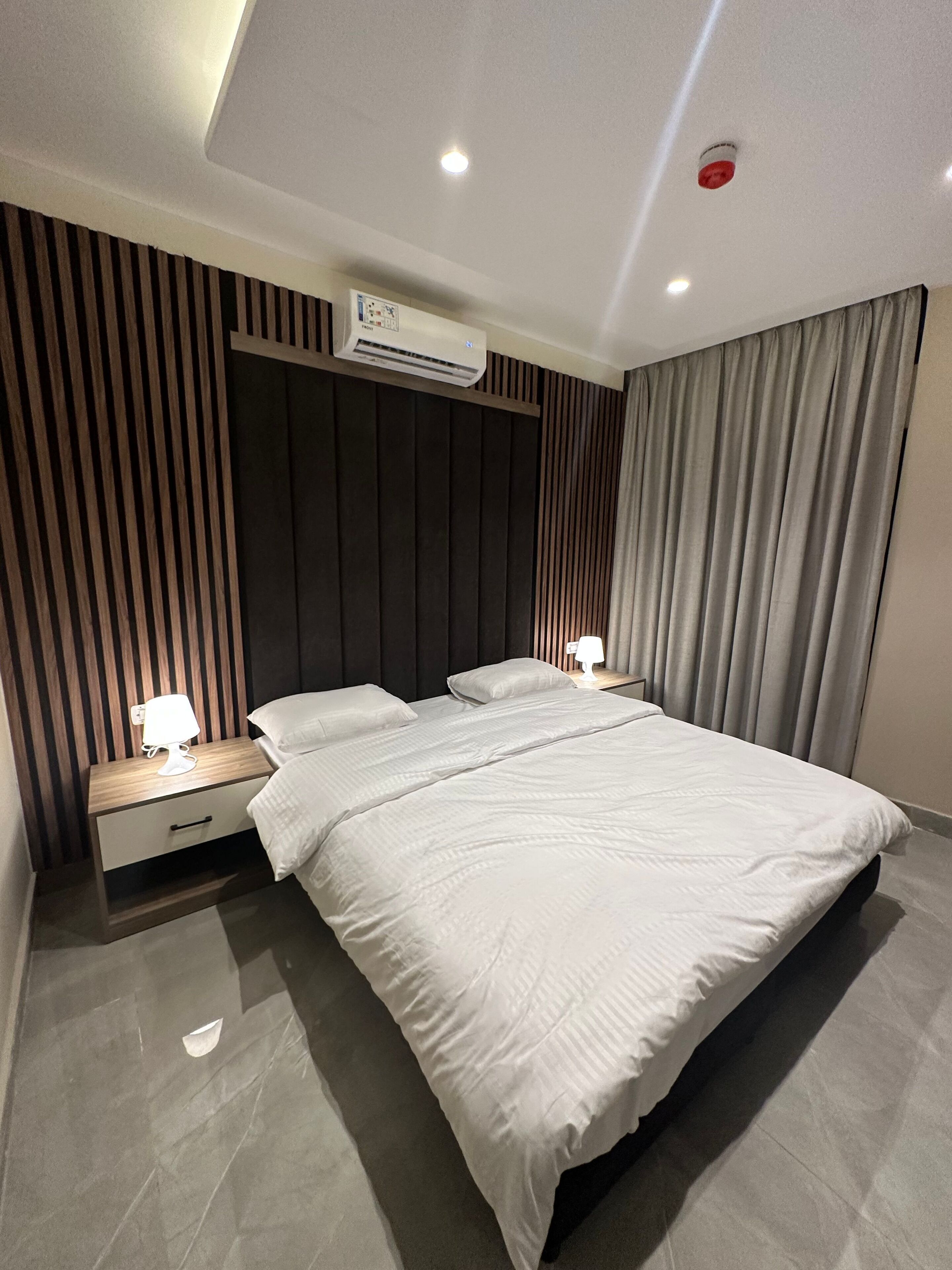 A modern hotel room with a large bed, a bedside table with a lamp, a wall-mounted air conditioner, and a ceiling light.