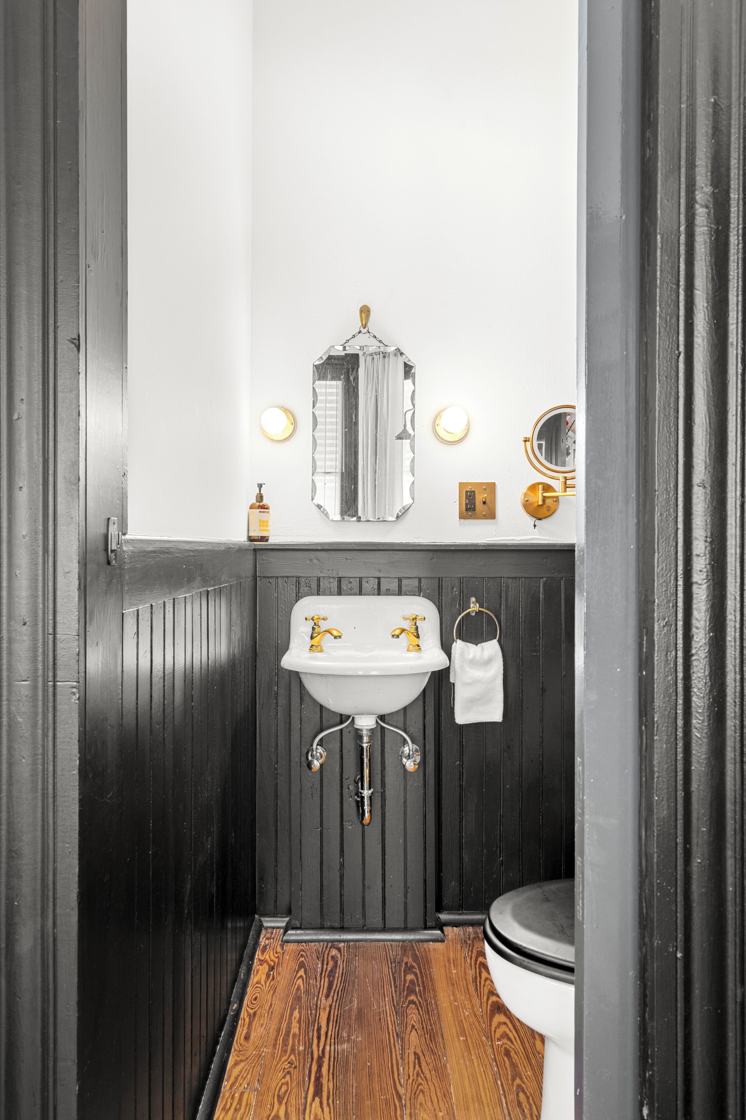 A bathroom with a wooden floor, a white sink with gold fixtures, a mirror, and a toilet.