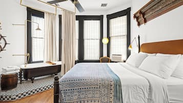 Camera d'albergo con un letto grande, una testiera in legno, una scrivania con una sedia, una finestra con tende, una ventola a soffitto e un pavimento a scacchi.