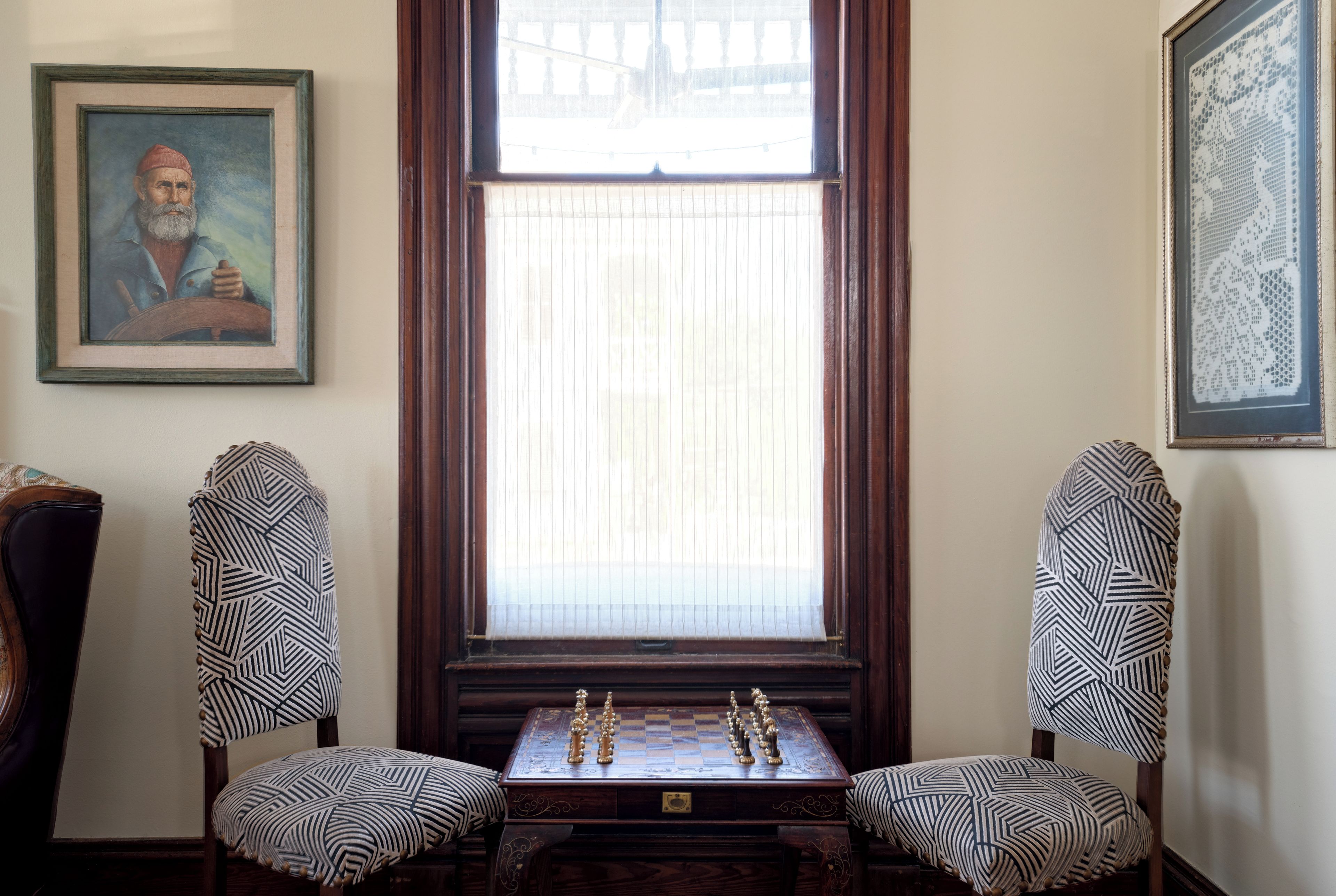 Une pièce comprenant un échiquier, deux fauteuils à motifs, un portrait encadré et une fenêtre avec des stores.