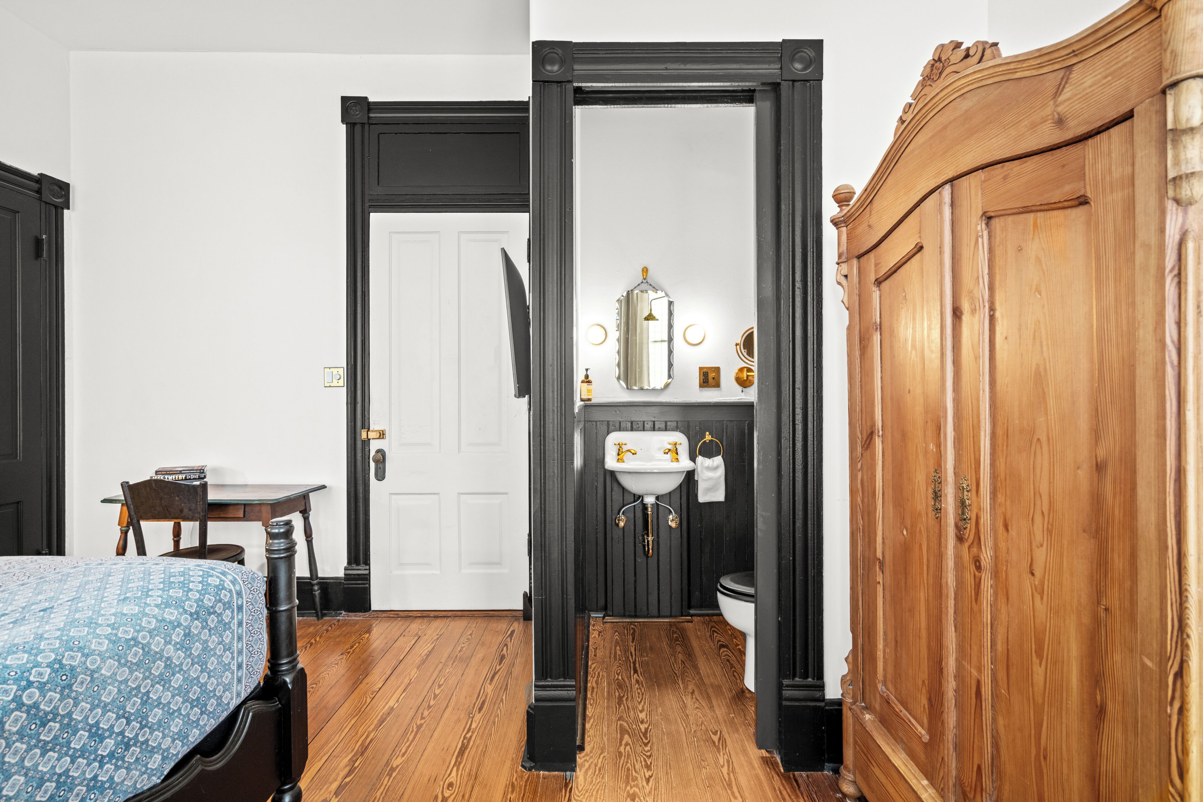 Une pièce avec un plancher en bois, une porte blanche, une armoire en bois, une petite table avec une chaise et un lit recouvert d’une couvre-lit à motifs.