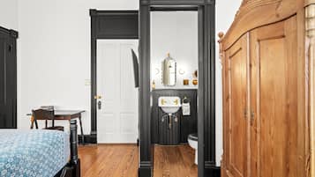 Une pièce avec un plancher en bois, une porte blanche, une armoire en bois, une petite table avec une chaise et un lit recouvert d’une couvre-lit à motifs.