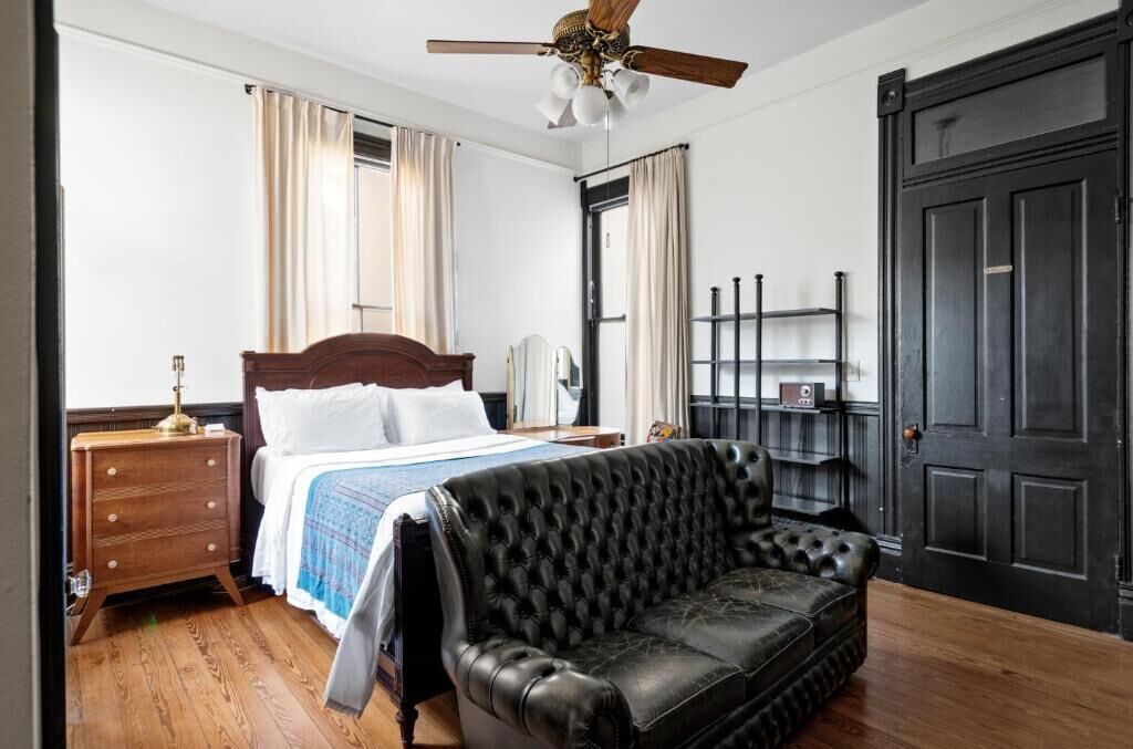 A bedroom with a bed, a black leather sofa, a wooden nightstand, a ceiling fan, and a black door.