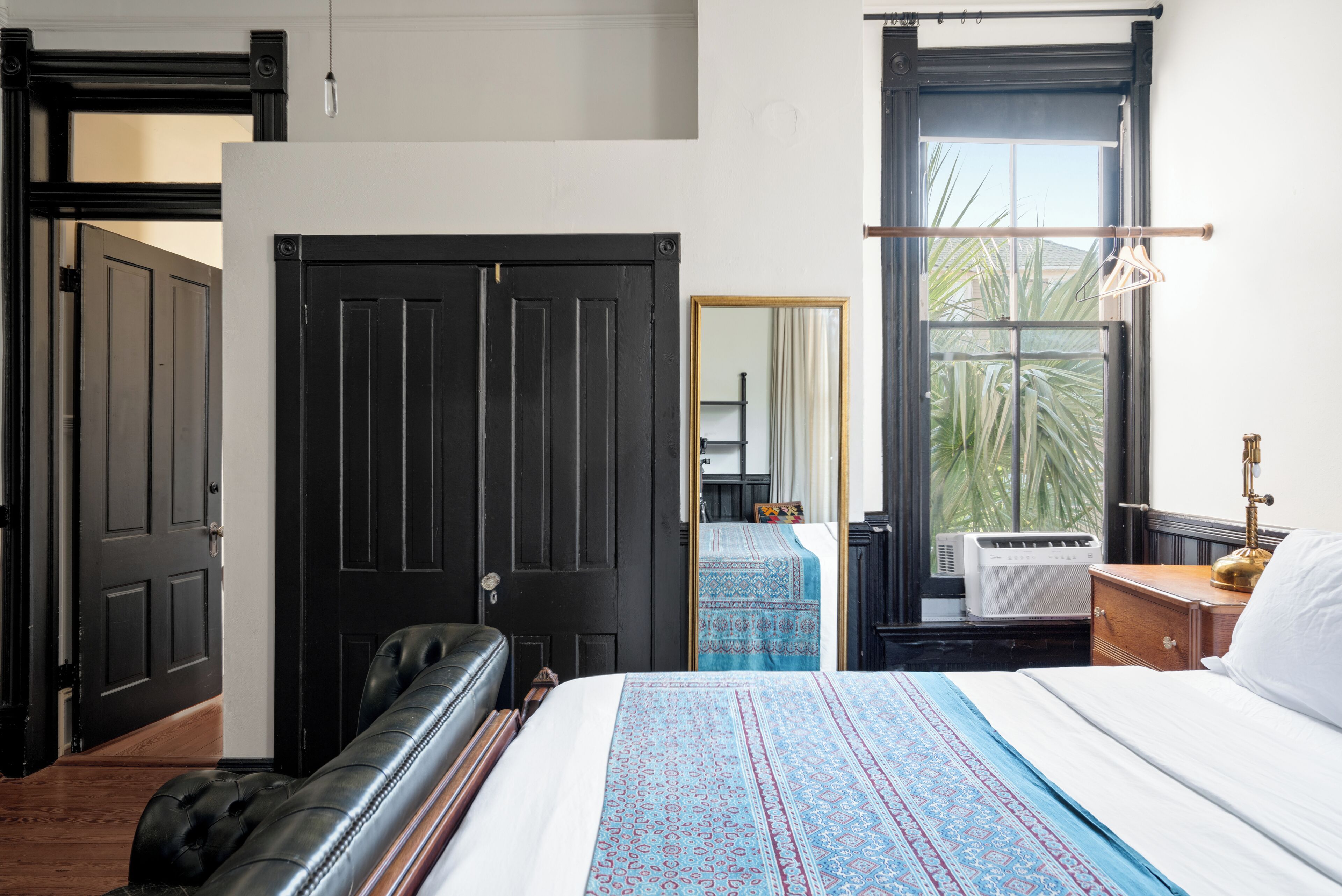 A bedroom with a large bed, a black leather sofa, a wardrobe, and a window with a view of palm trees.