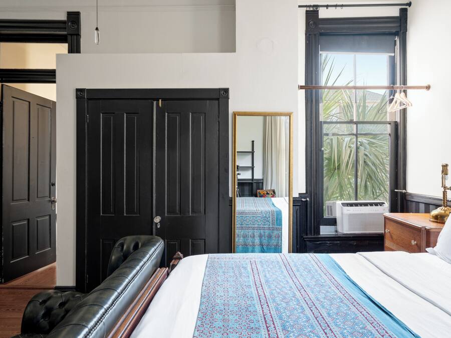 A bedroom with a large bed, a black leather sofa, a wardrobe, and a window with a view of palm trees.
