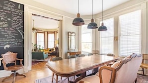 Uma sala de jantar com uma grande mesa de madeira, cadeiras estofadas, iluminação pendente e um quadro negro com escrita.
