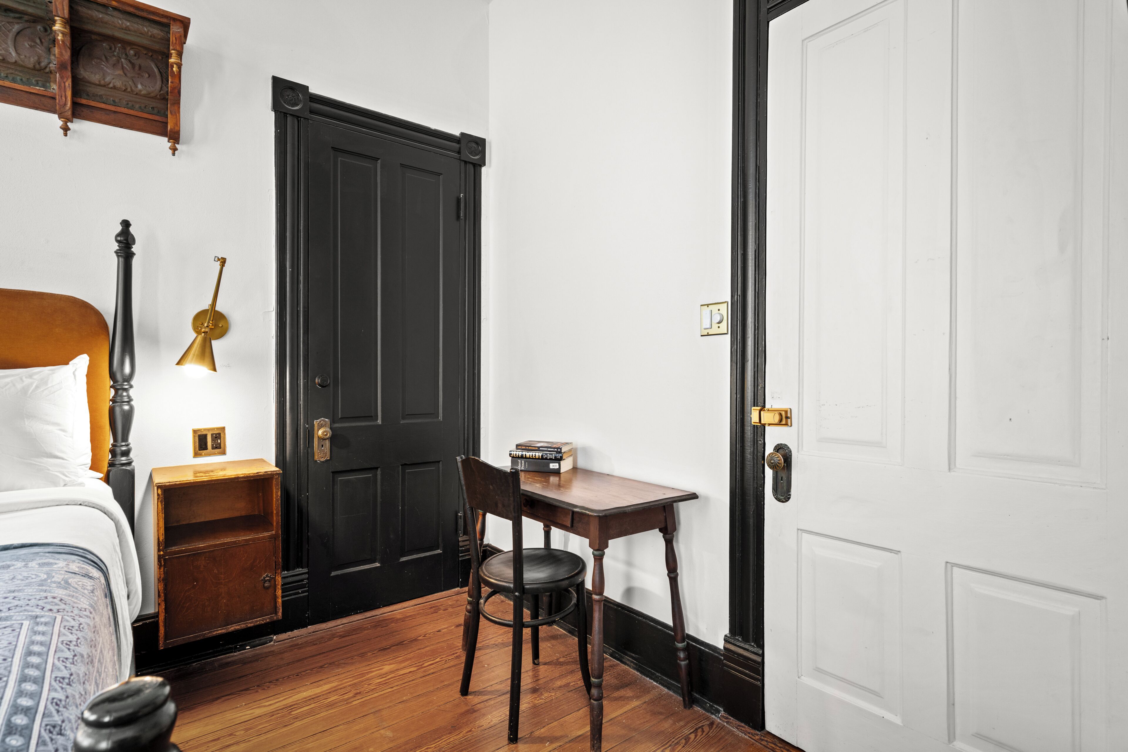 A room with a bed, a nightstand, a desk with a chair, a black door, and a white door.