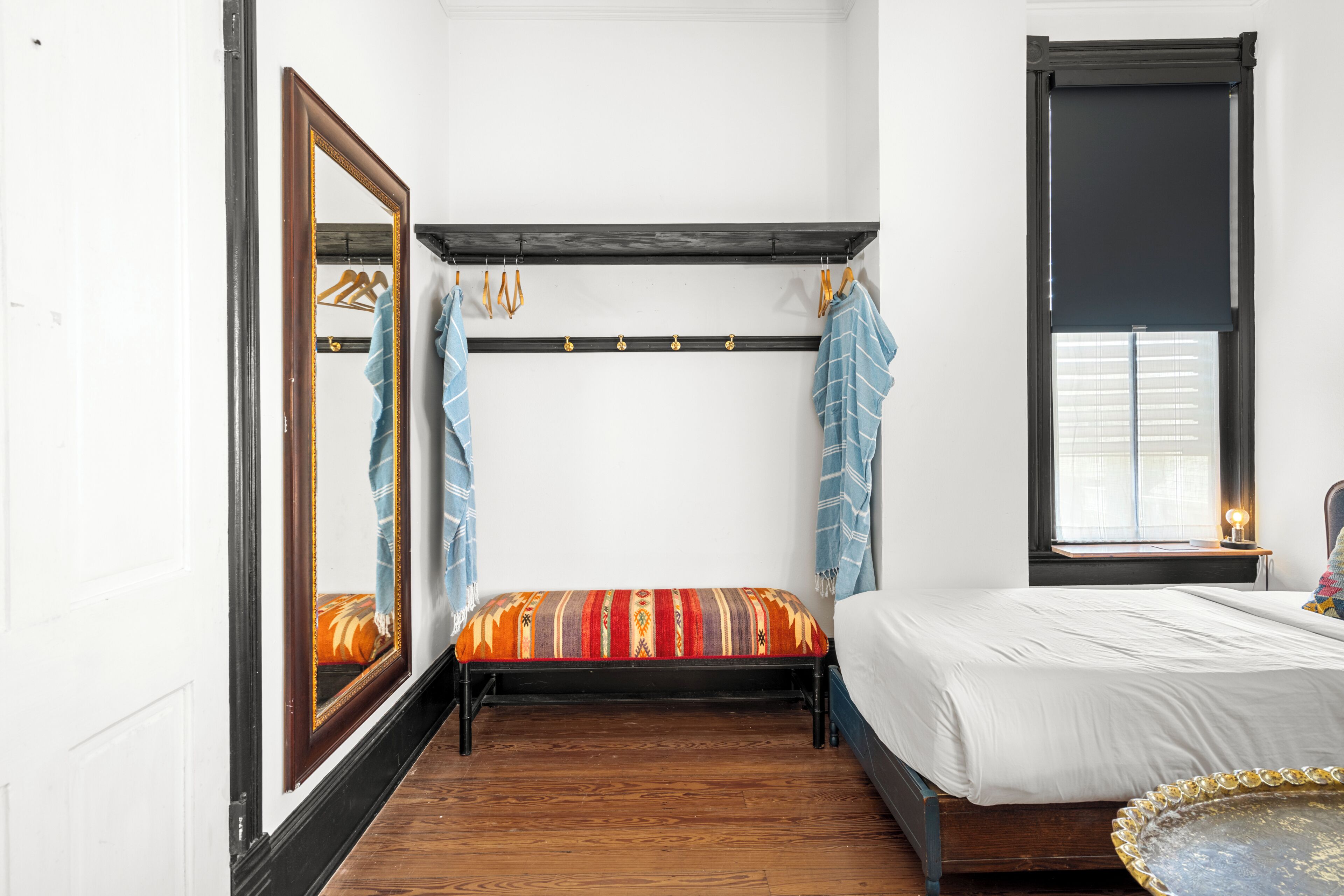 A room with a bed, a large mirror, a window with blinds, and a wooden floor.