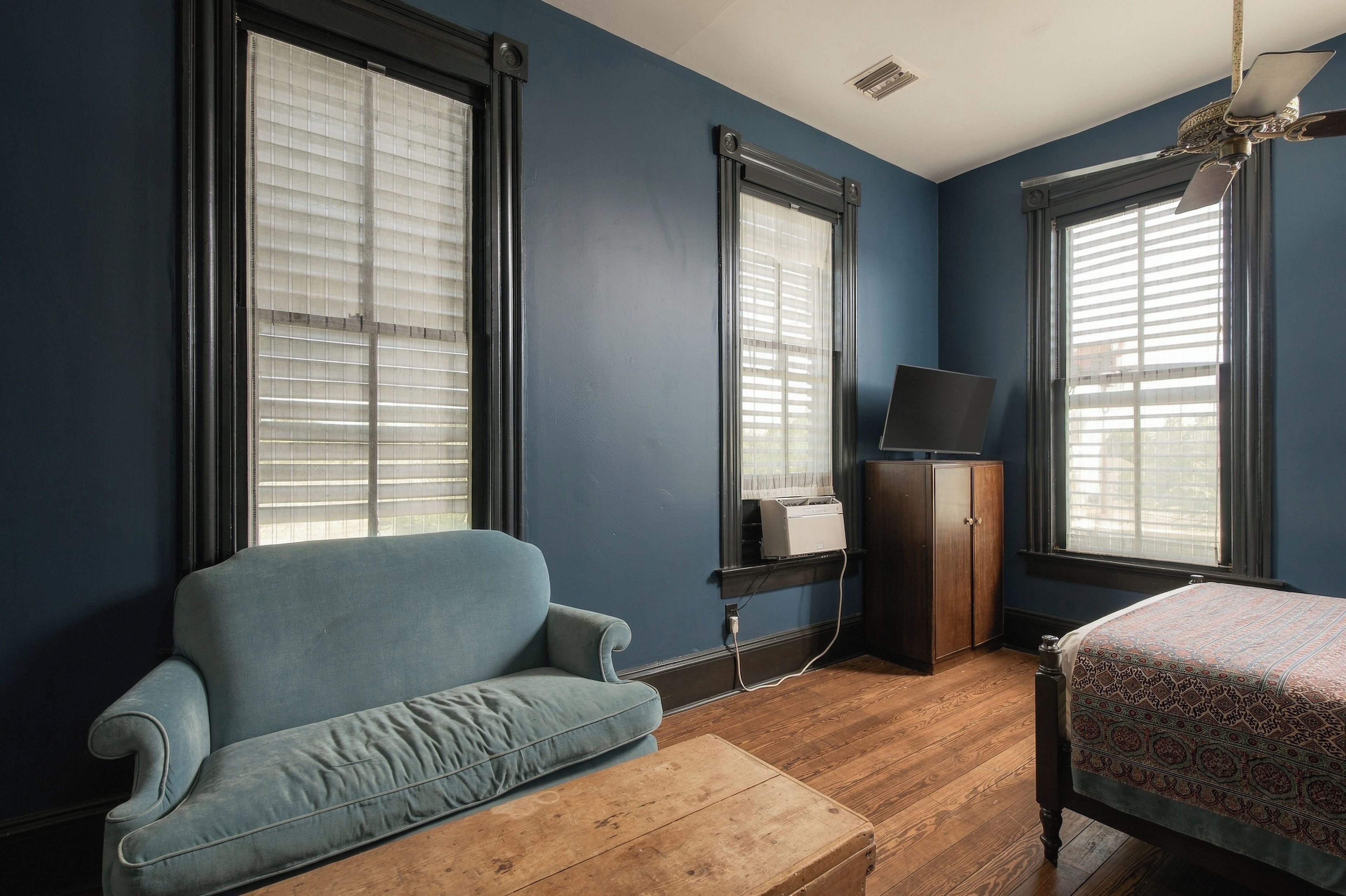 A room with a blue wall, a wooden floor, a blue sofa, a bed with a patterned bedspread, and a wooden cabinet.